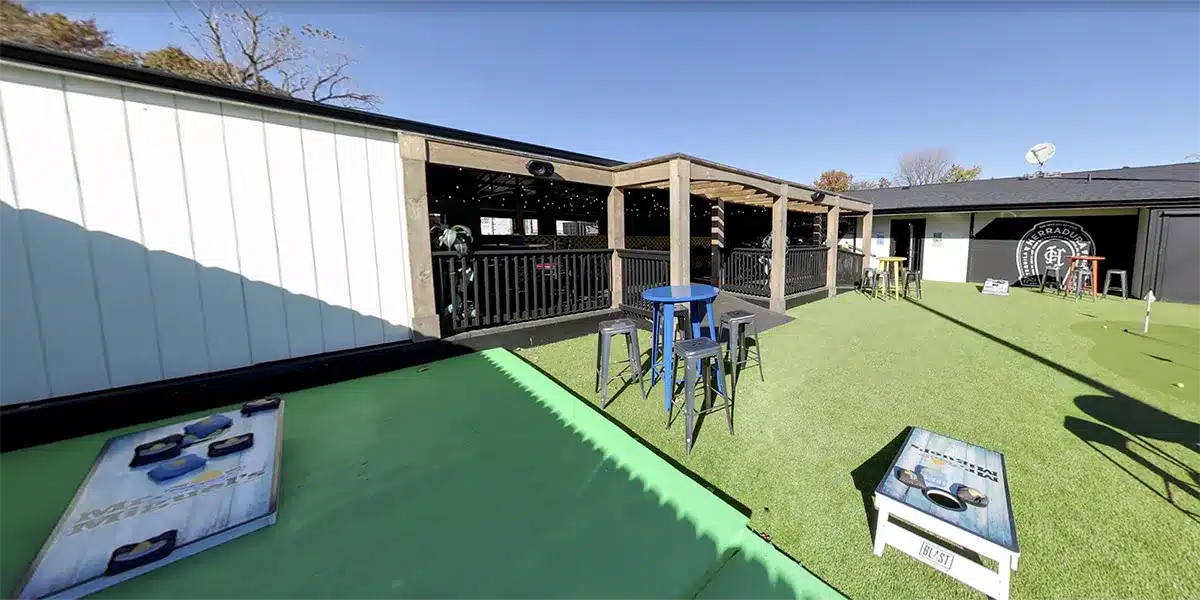 Outdoor space featuring green turf, tables, and a covered seating area beside a building under a clear blue sky.